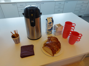 Kuvassa valkoisella pöydällä pullapitko, kahvimukeja ja termospullo, jossa kahvia.